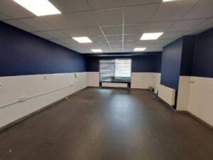 An interior photograph of a large, empty room. The walls are painted navy blue on the top half, with white tiled blockwork on the bottom half. The room features a dark grey floor, a suspended grid ceiling with square LED panel lights, and a window with dark blinds at the far end. White radiators are positioned beneath the window and along the right-hand wall, and electrical trunking runs along the left wall.