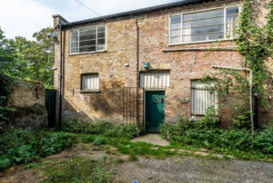 An exterior shot of a two-story aged brick building featuring a central dark green door, barred ground-floor windows, large multi-pane upper windows, and a courtyard filled with overgrown weeds and climbing ivy.