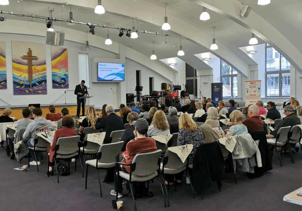 A man in a suit speaks at a podium in front of a seated audience at the Thanet District Council annual landlord event. A large screen behind him shows the event title. People are seated in rows facing the front, with some tables visible at the sides. Musical instruments and equipment are set up to the right of the stage. Colorful tapestries hang on the wall behind the podium.