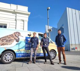 Colourful birdwise transit van sits in front of bright blue sky. Three people standing in front smiling.