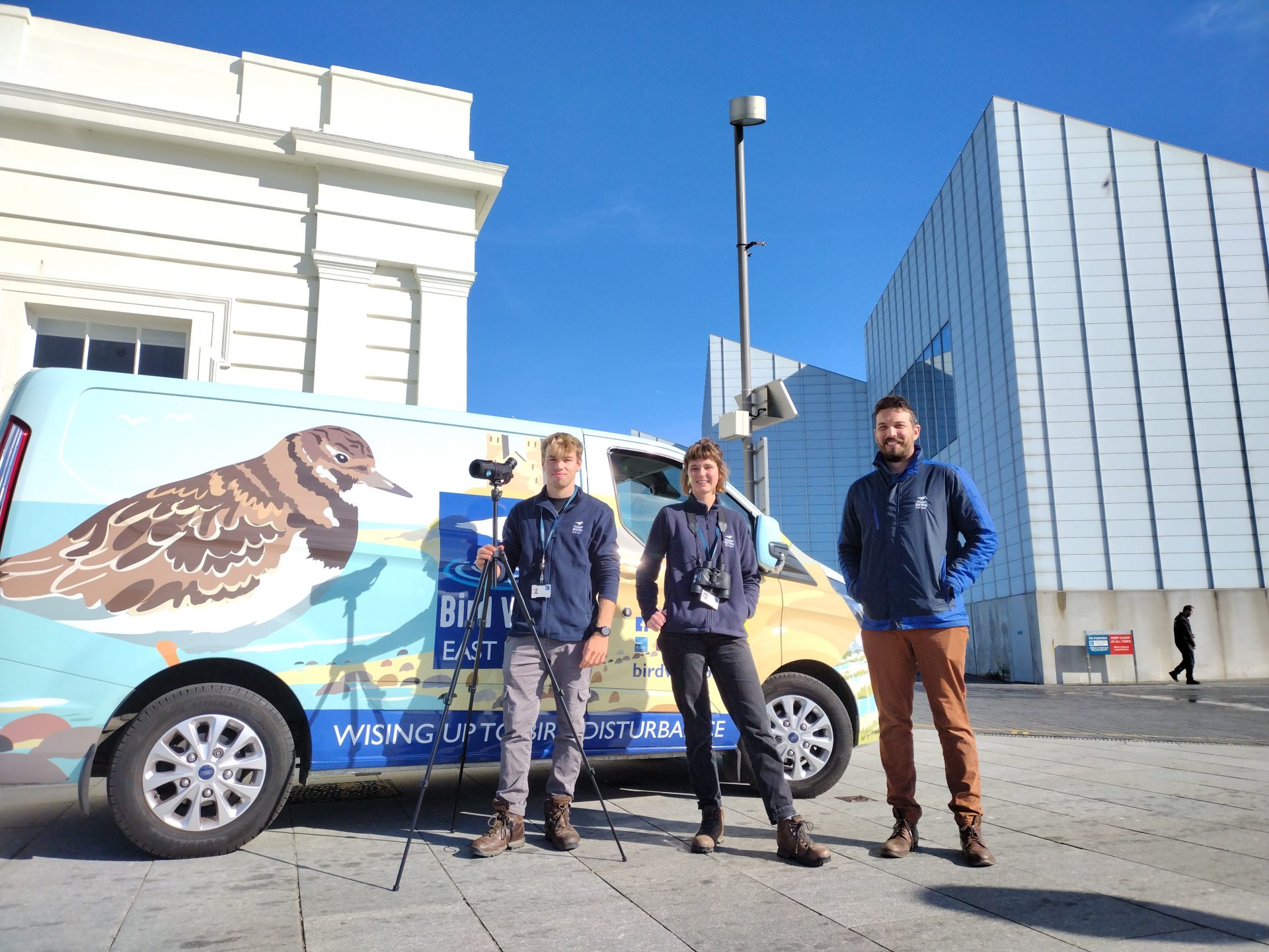 Colourful birdwise transit van sits in front of bright blue sky. Three people standing in front smiling.