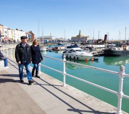 Two people walking on the left hand side adjacent to railings and blue sea. Moored boats and buildings in the background of the photo.