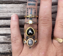 photo of a white skinned hand on a light wood background. Ring finger is tattooed and has five different rings on it.