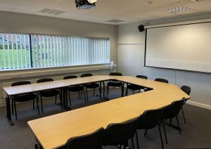 A set of tables and chairs organised in a c shape with a projector screen