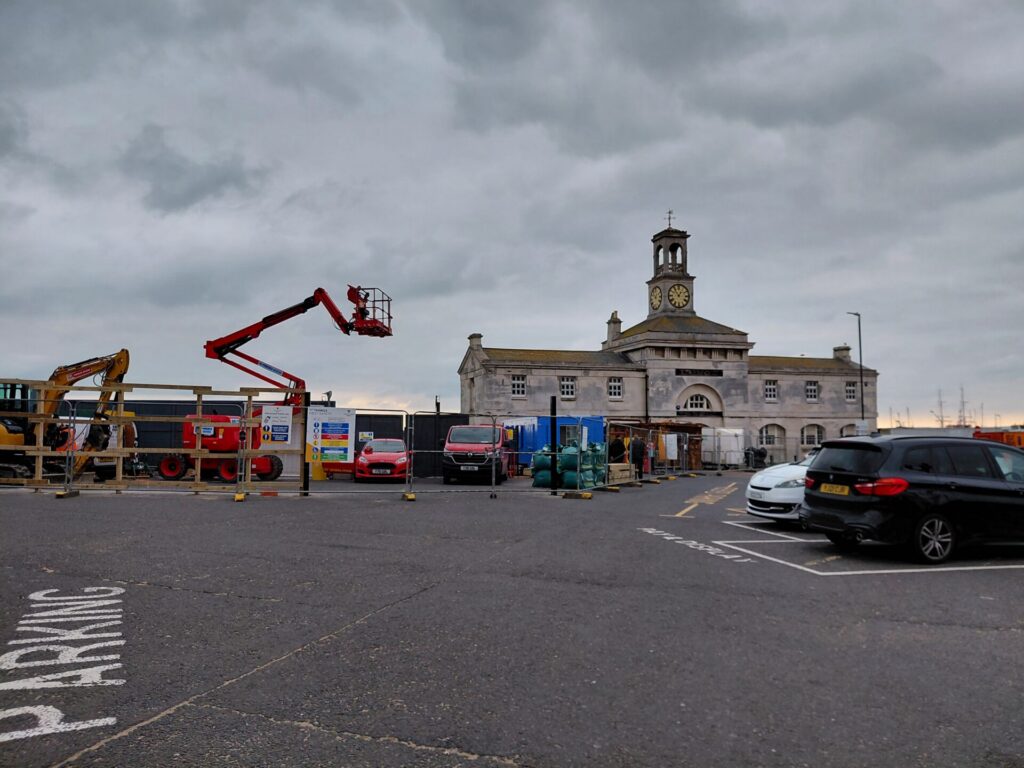 An exterior shot of the historic Clock House from across a parking lot under an overcast sky. The site is surrounded by construction fencing, with an elevated red cherry picker and a yellow excavator positioned next to the building.