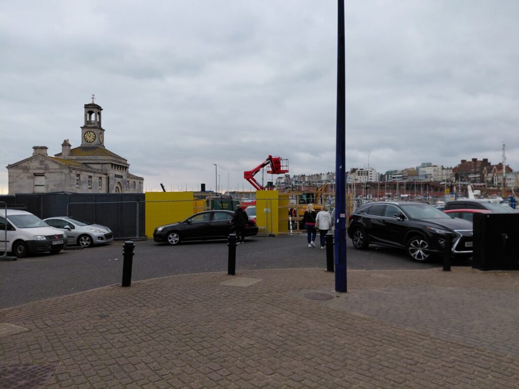 An exterior view of the Clock House and Ramsgate harbour from a nearby car park on an overcast day. Yellow construction hoarding surrounds the building's base, with a red cherry picker visible behind it, and several parked cars in the foreground.