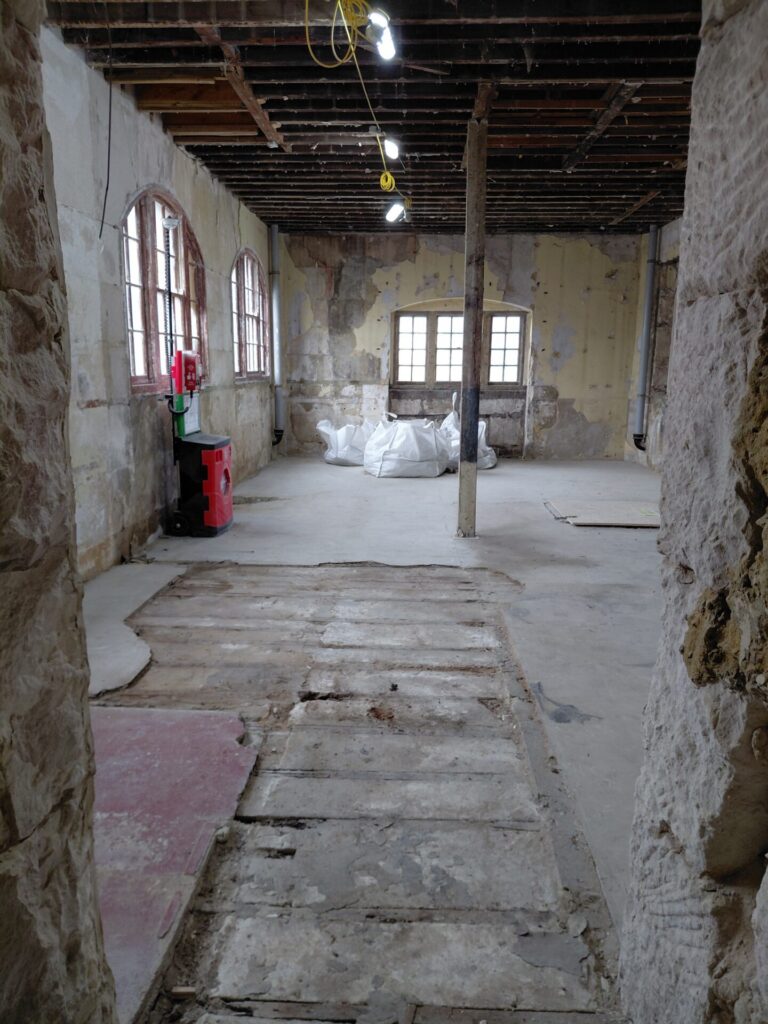 Interior view of a stripped-back room with exposed ceiling joists, bare walls, and temporary lighting. Several large white rubble sacks sit in the center of the floor near the windows, and a red mobile fire safety point stands on the left.