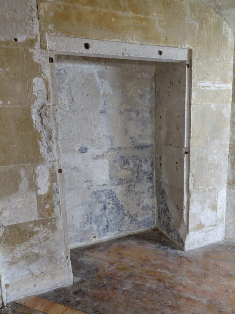 Close-up of a blocked-in rectangular alcove or former doorway in a bare interior wall. The wall shows layers of scraped plaster and peeling paint, with exposed wooden floorboards at the base.