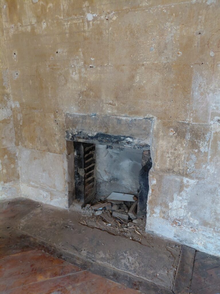 Close-up of a hollow, stripped-out fireplace opening containing broken rubble, set into a bare, unfinished wall above exposed wooden floorboards.