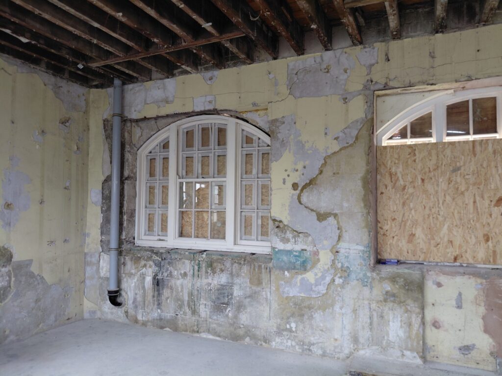 A distressed interior wall with peeling yellow and blue paint, featuring a large arched wooden window in the center. Some of the window panes are boarded up with wood, and the ceiling above has exposed wooden joists.