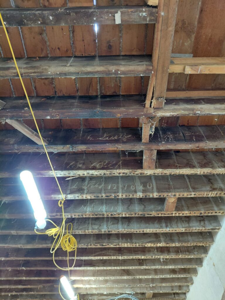 Looking up at an exposed wooden ceiling inside the Clock House, showing old wooden joists with faded historical writing on them, illuminated by a temporary construction light.