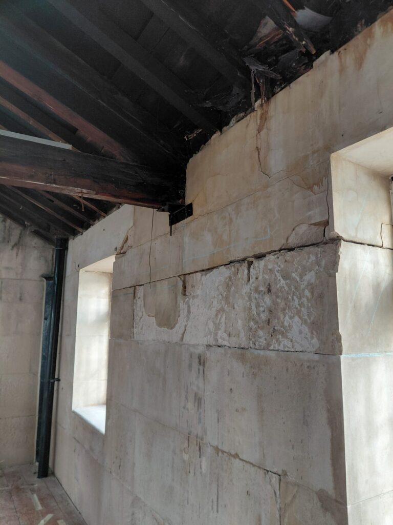 Close-up of an interior corner showing thick stone block walls meeting a dark, exposed timber roof structure. There are visible cracks in the stonework, faint blue construction markings, and a black pipe running vertically down the wall.