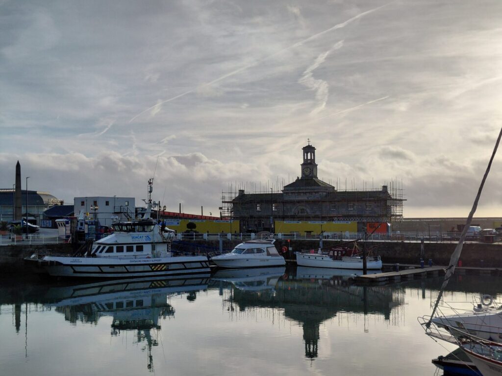 The Ramsgate Clock House covered in scaffolding, viewed across the harbour with several boats moored in the foreground and the building's reflection in the water.