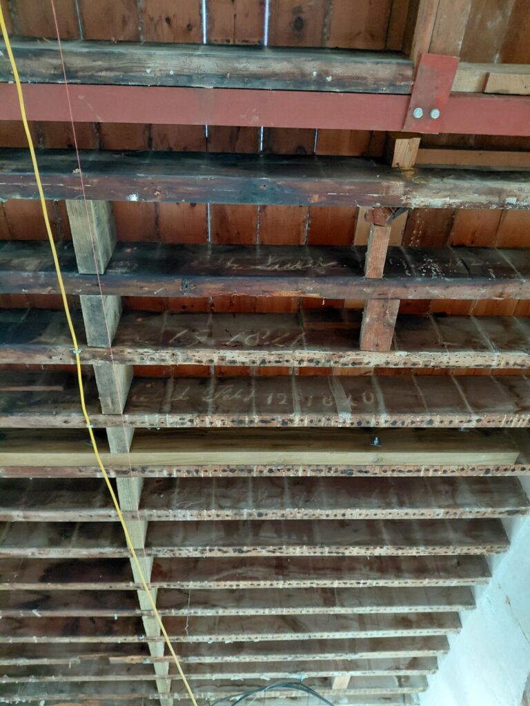 Close-up of the exposed wooden ceiling structure inside the Clock House, showing historic floor joists with faded handwritten dates and script, alongside a modern red steel support beam and temporary wiring.