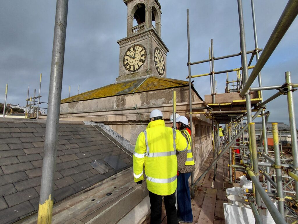 Two construction workers in hard hats and high-visibility jackets standing on roof-level scaffolding, inspecting the slate roof and the historic stone clock tower.