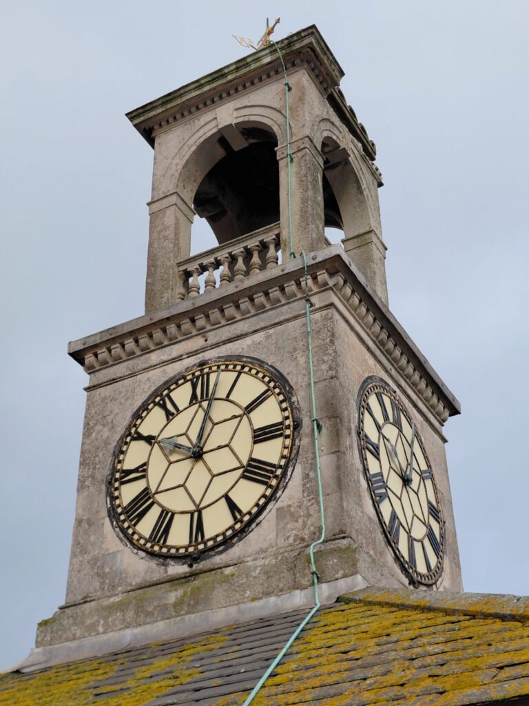 A close-up looking up at the historic stone clock tower under a grey sky. It features a bell cupola at the top, two clock faces with Roman numerals, and a green lightning conductor wire running down its side.