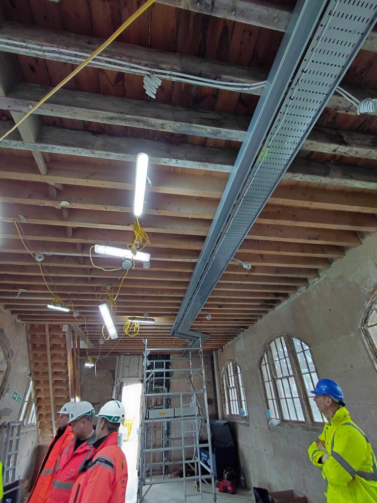 Interior view showing construction workers in high-visibility gear standing under an exposed wooden ceiling with new metal cable trays, temporary lights, and large arched windows.