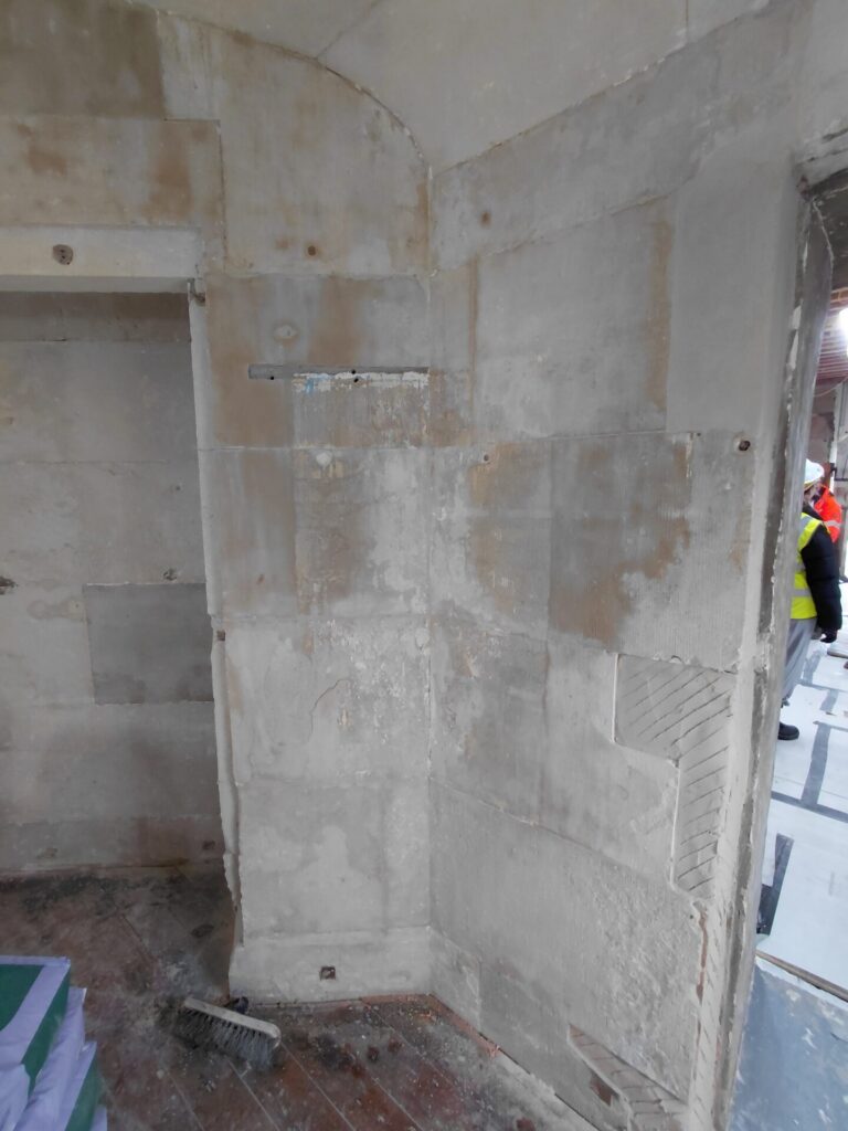 An interior stone wall undergoing renovation inside the Clock House, with a doorway on the left and a worker partially visible in the background.