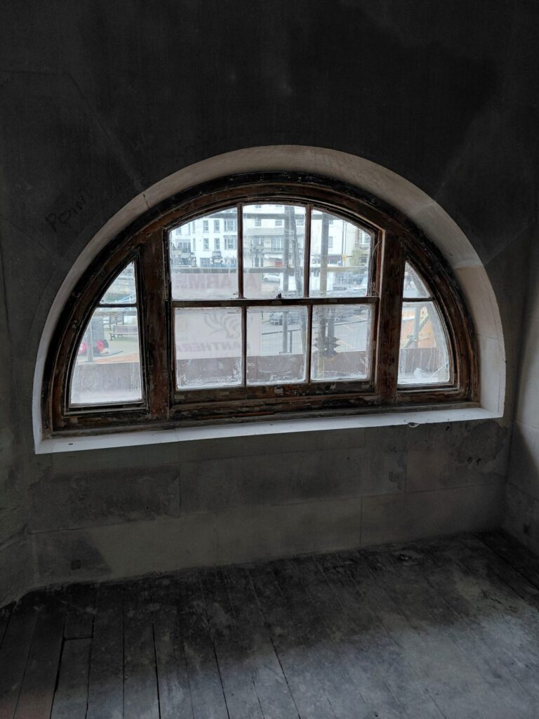Looking from the inside of a dark, unfinished room out through a large, arched wooden window. Scaffolding and a neighboring building are faintly visible through the dusty glass.
