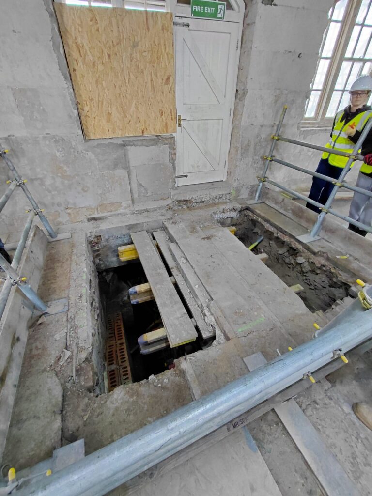 An interior floor under construction, featuring a large, deep rectangular trench opened up in the ground. Temporary wooden planks lie across the gap, surrounded by metal scaffolding guardrails. A worker in high-visibility gear stands to the right.