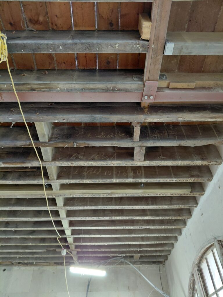 Looking up at the exposed wooden ceiling structure inside the Clock House, showing historic floor joists with faded handwritten dates and text, alongside modern temporary lighting.