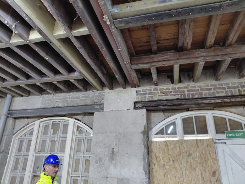 Interior view showing exposed wooden ceiling joists reinforced with modern steel beams, above a stone wall with large arched windows. A construction worker in a blue hard hat is partially visible in the bottom left.