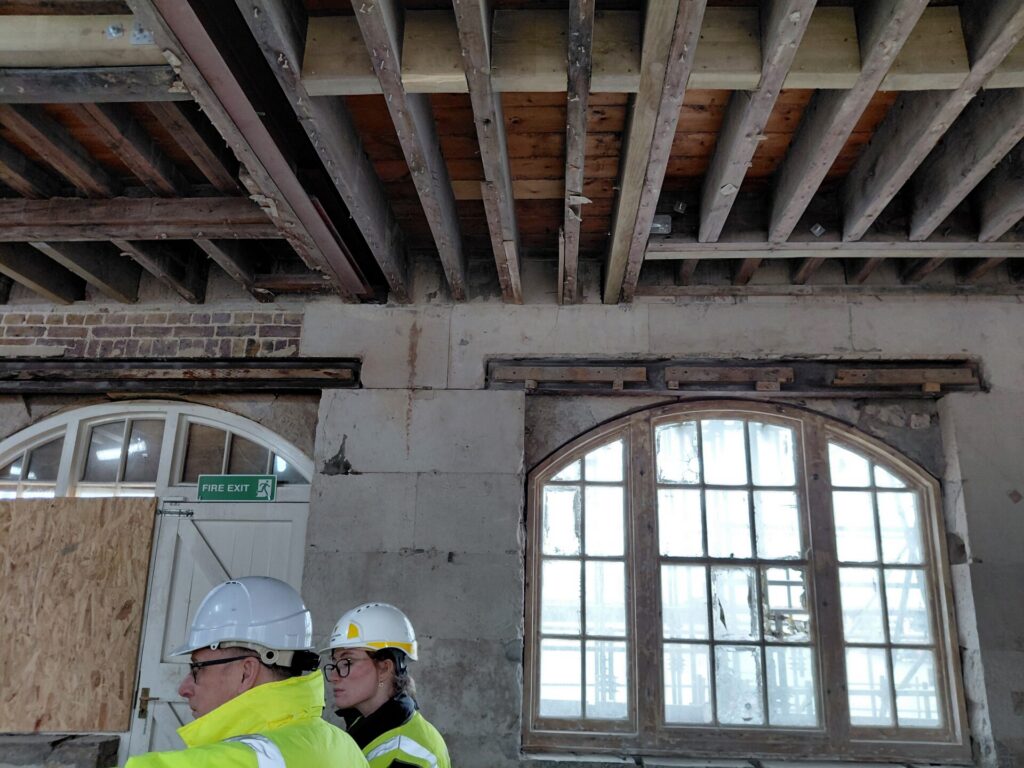 Two people in hard hats and high-visibility jackets standing inside the Clock House during renovations, with exposed ceiling beams and large arched windows behind them.
