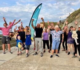 Large group of mixed age and gender people and trainer posing for a photo on the lower promenade in front of a proportional feather flag with hands in the air cheering