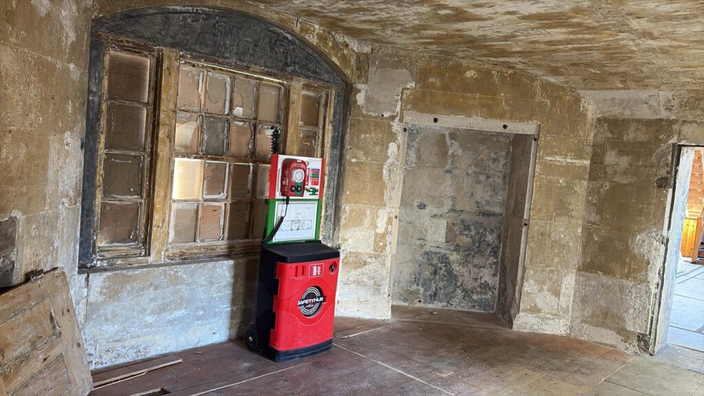 A stripped-back interior room with bare stone walls and a large, old multi-paned wooden window. A bright red mobile fire safety and first aid point stands in the center of the dusty wooden floor.