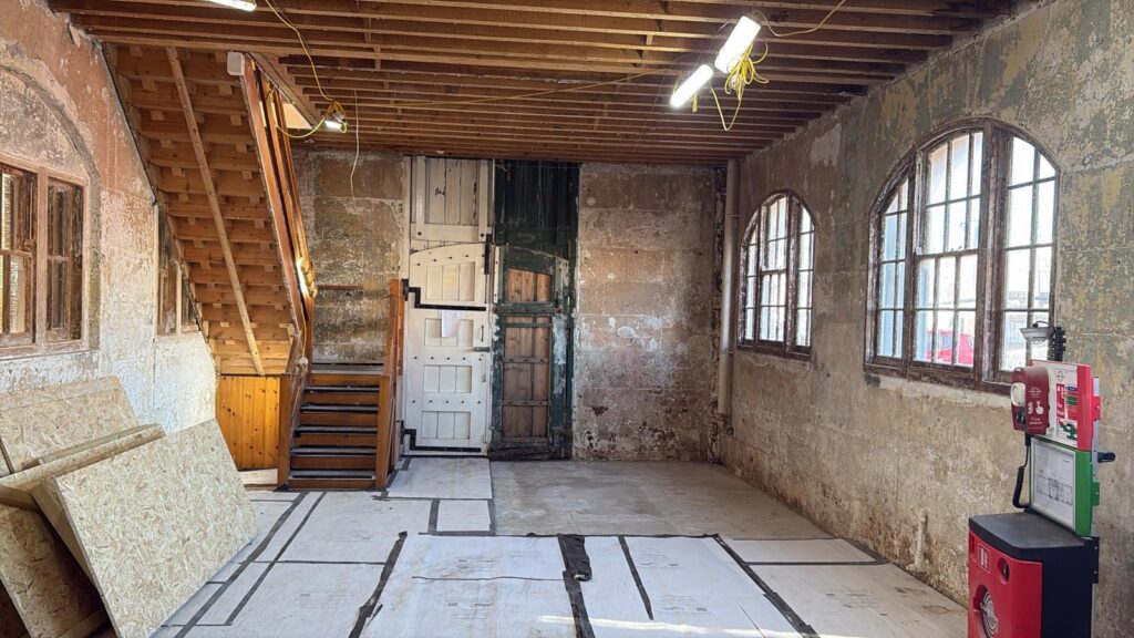 Interior view of a renovation space with exposed ceiling joists and temporary lights. The room features a wooden staircase on the left, tall historic wooden doors in the center, and two arched windows on the right next to a fire safety point.