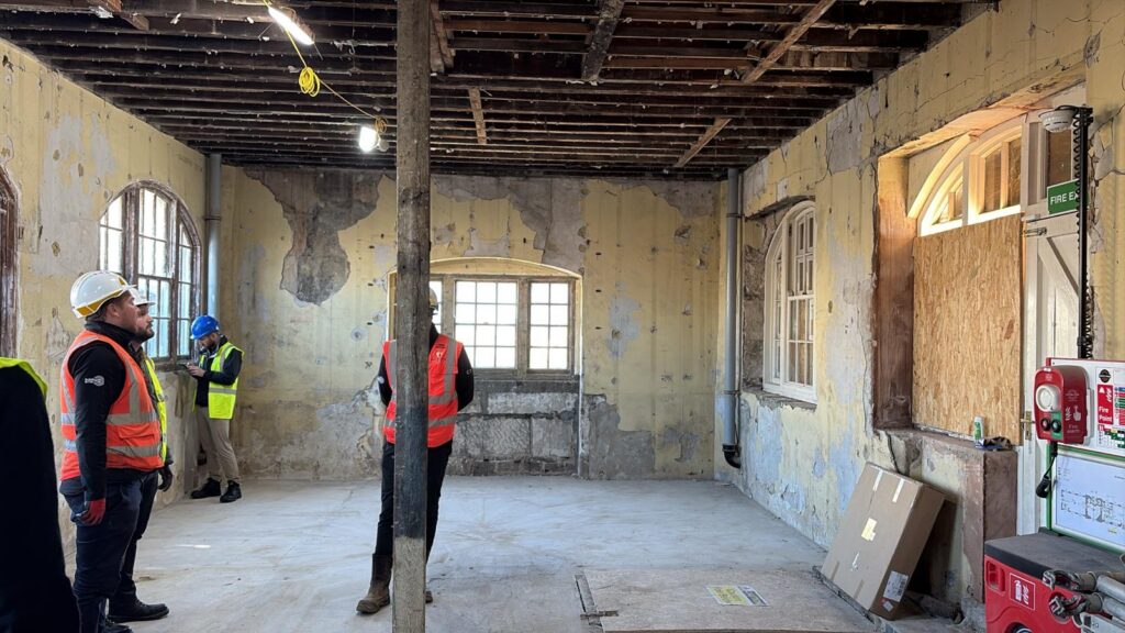 Three people in hard hats and high-visibility vests stand conversing inside a large, stripped-back room. The space has exposed ceiling joists, bare walls with peeling paint, arched windows, and a central wooden support pillar.