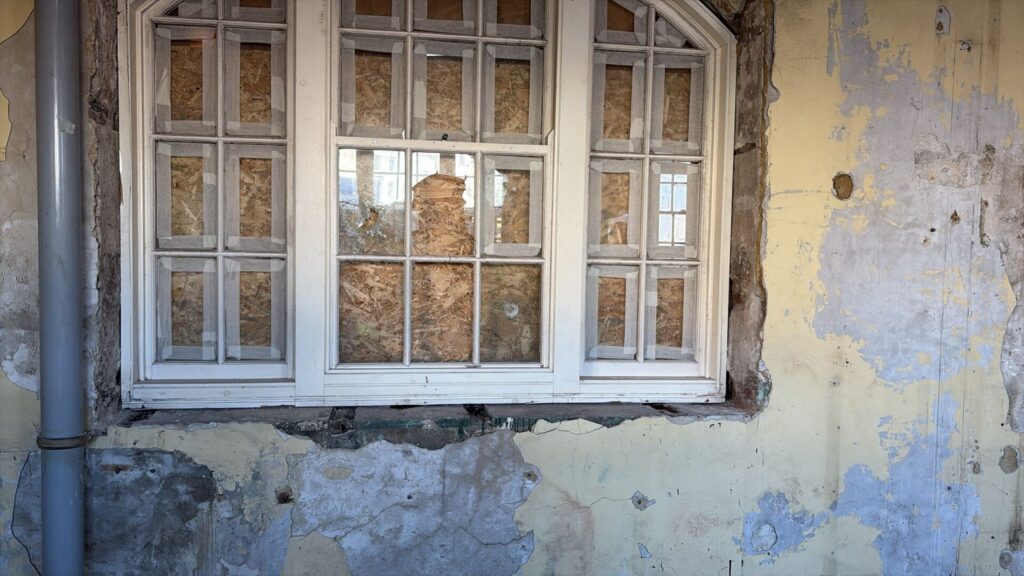 Close-up of a historic white wooden window frame. The glass panes are taped and boarded up with wood from the inside, set into a wall with peeling yellow paint and exposed underlying plaster.