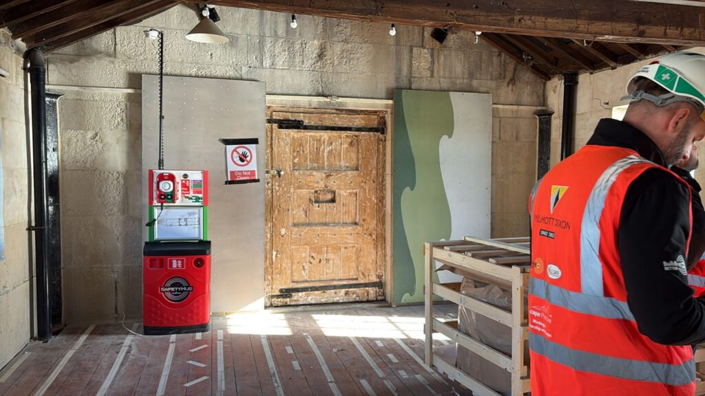 An upper-level room with an exposed timber roof and a sturdy historic wooden door in the center. A red mobile fire safety point is on the left, and a construction worker in an orange Willmott Dixon high-visibility vest stands on the right.