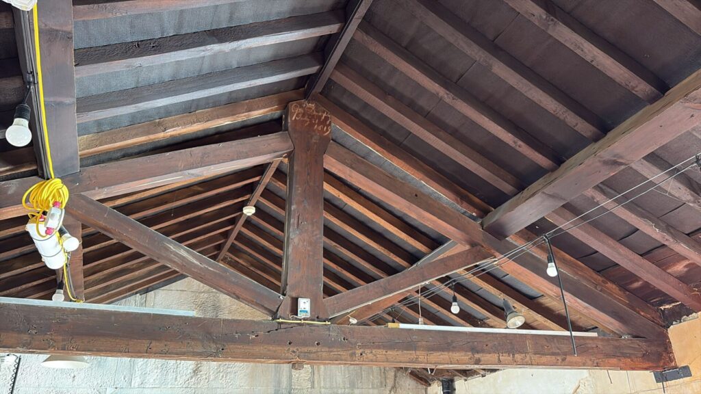 Close-up of the exposed, dark timber roof trusses and beams inside the building. Faint historical handwriting is visible carved into a central wooden post, alongside modern temporary string lighting.