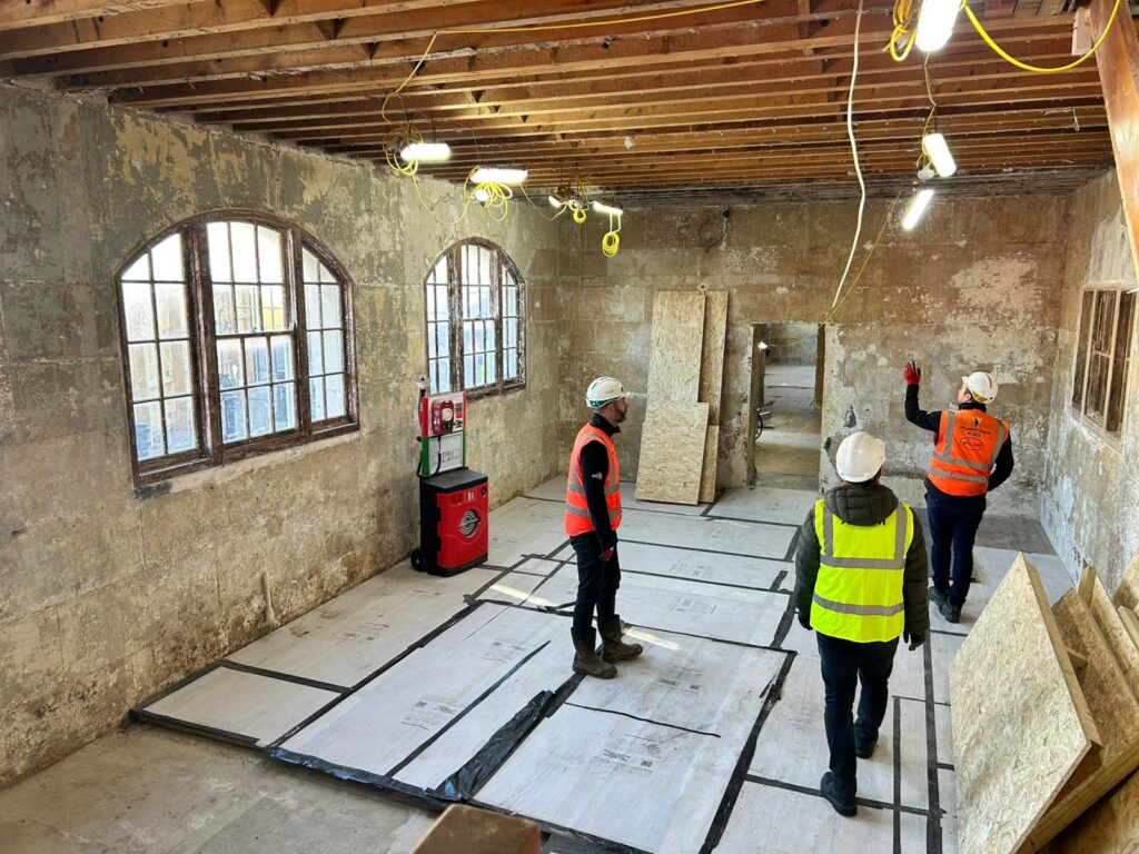 Three people in hard hats and high-visibility gear inspecting an unfinished room. The floor is covered in taped protective sheeting, and temporary lights hang from the exposed ceiling joists. One worker is pointing at the bare, distressed plaster wall on the right.