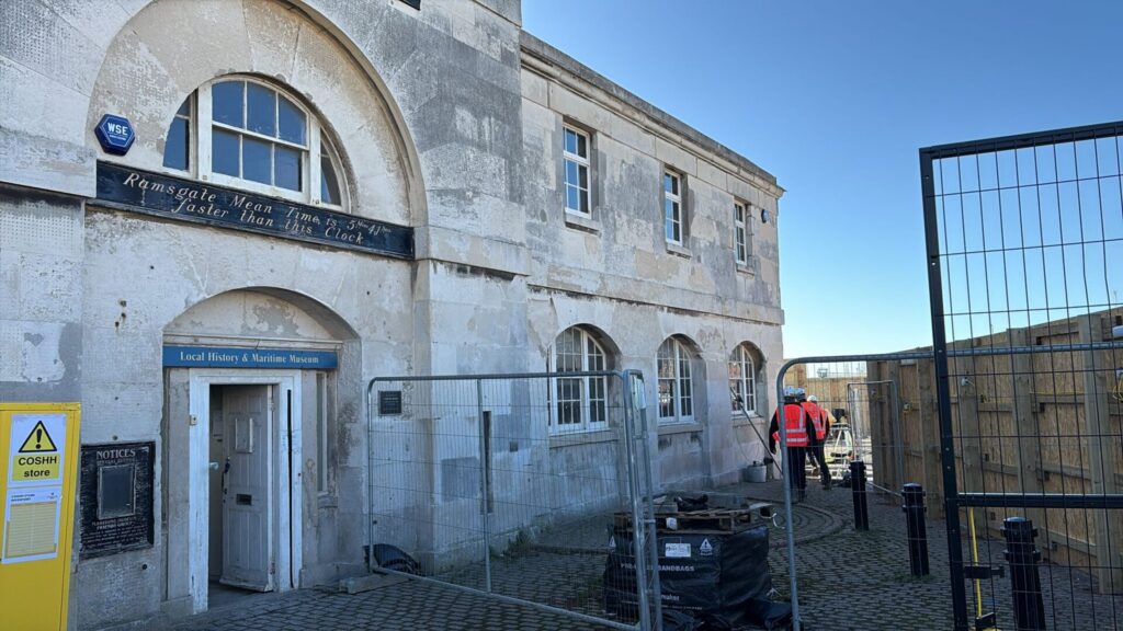 Exterior of the historic stone Clock House building under a clear blue sky. Signs above the entrance read 'Local History & Maritime Museum' and 'Ramsgate Mean Time is 5 Mins 41 Secs faster than this Clock'. Metal construction fencing and a yellow safety cabinet sit in front, with two workers in high-visibility gear walking on the right.
