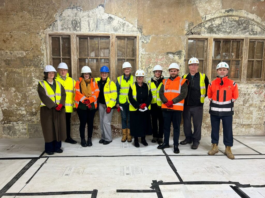 A group of ten people wearing hard hats and high-visibility jackets smiling for a photo inside the building. They are standing on a floor covered in protective white boarding, in front of a distressed stone wall and a large, multi-paned wooden window frame.