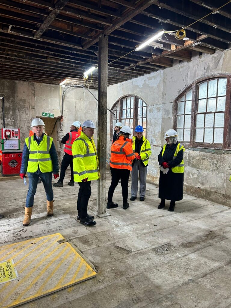 An interior room during renovations, showing exposed ceiling joists and large arched windows. Several people in hard hats and high-visibility vests are standing in small groups talking, with a red mobile safety hub visible on the left and a yellow caution cover over a hole in the floor.