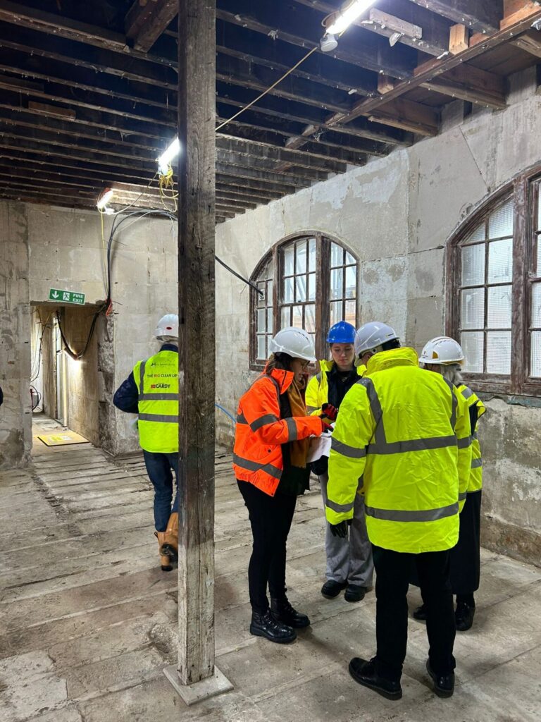 A group of four people in hard hats and high-visibility jackets gathered together in a stripped-back room, reviewing documents. Another worker walks away down a corridor toward a green fire exit sign in the background.