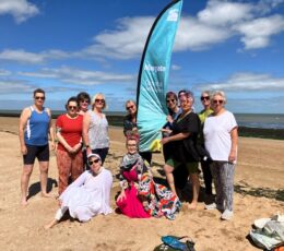 Group of people on the beach with the sea in the background posing for photo next to event sail banner on a sunny day