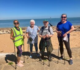 Four adults with poles standing on lower promenade posing for photo in front of sand beach during their nordic walk