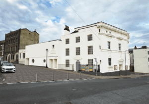 An architectural rendering of the proposed restoration for the Theatre Royal Margate. The main building features a pristine, restored white facade. To the left, a new modern extension has been added with white vertical cladding and two circular windows. The street front has been transformed into a paved public area with grey tiling and bollards.