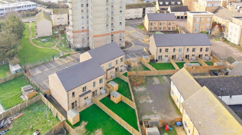 Aerial view of a new housing development featuring two blocks of modern, light-brick terraced houses with dark roofs. The homes have freshly laid, fenced rear gardens with small wooden sheds, and sit next to newly paved parking areas within an existing residential neighbourhood.