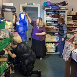 A store room of colourful folded fabrics, trays and crates of materials. Spools of yarn oar on a shelf. Three people are looking through the stock. One of them is placing a tiara on the head of a manakin swathed in gauzy purple fabric.