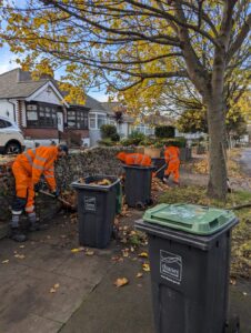 Three people dressed in orange high visibility protective clothing are busily collecting fallen leaves from a residential street. There is a large tree nearby and autumnal leaves litter the ground.