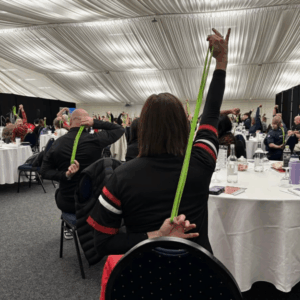 A view from behind of conference attendees participating in a seated movement break inside a large marquee. The room is filled with round tables, and participants are stretching using green resistance bands, with one arm reaching up and the other reaching behind their back.