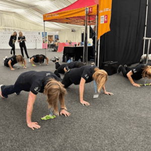 A group of team members wearing matching dark t-shirts perform push-ups on the floor of the event hall. In the background, two colleagues stand and smile, while a 'Kent County Council' banner hangs on the right.