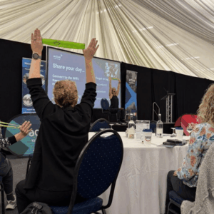 A view from behind of a conference attendee raising a green resistance band overhead with both hands. In the background, the presenter on stage mirrors the action, leading the group exercise in front of a large presentation screen.
