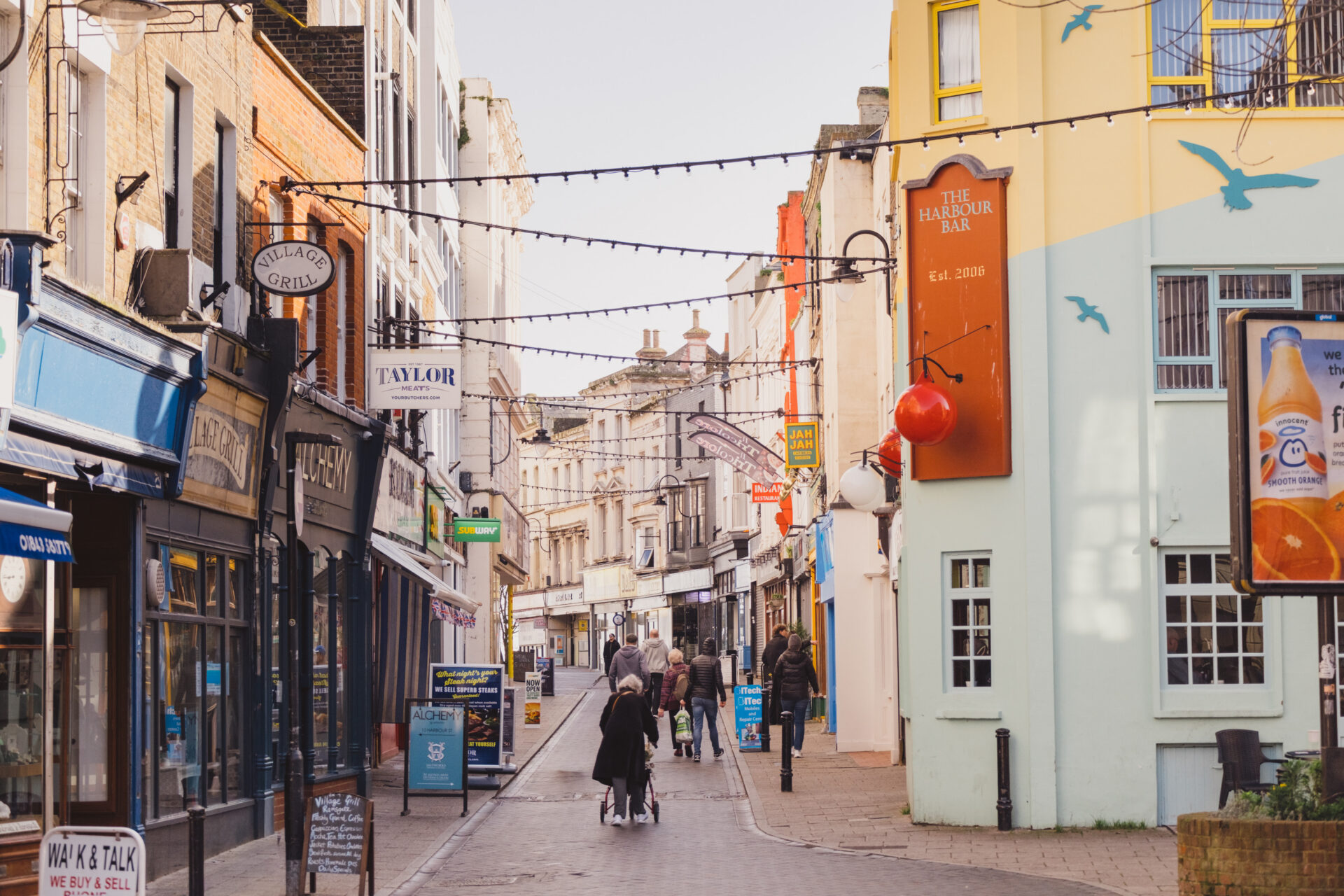 A sunlit, pedestrianised high street in Ramsgate lined with various shops and cafes. Strings of festoon lights zig-zag overhead across the road. On the left, shopfronts include Village Grill, Alchemy, and Taylor Meats. On the right stands a brightly painted yellow and light blue building featuring a large sign for 'The Harbour Bar' and painted seagulls. Several pedestrians are strolling down the street, including a person using a four-wheeled walker in the foreground.