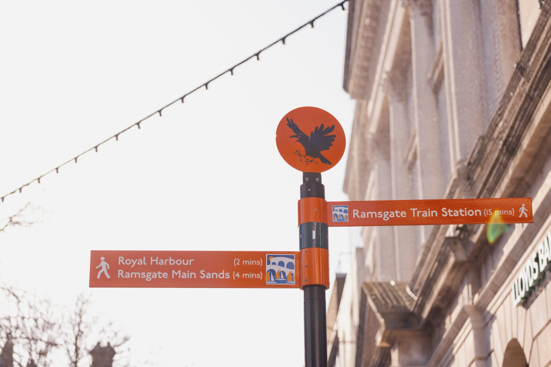 A bright orange pedestrian signpost in Ramsgate on a sunny day. The top of the post features a circular sign with a black bird silhouette. The left-pointing sign reads: 'Royal Harbour (2 mins)' and 'Ramsgate Main Sands (4 mins)'. The right-pointing sign reads: 'Ramsgate Train Station (15 mins)'. The background shows out-of-focus festoon lights and a classic stone building.